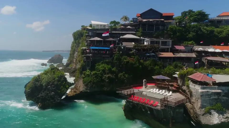 Aerial frame of a clifftop resort above turquoise surf, its terraces and umbrellas stepping down the headland