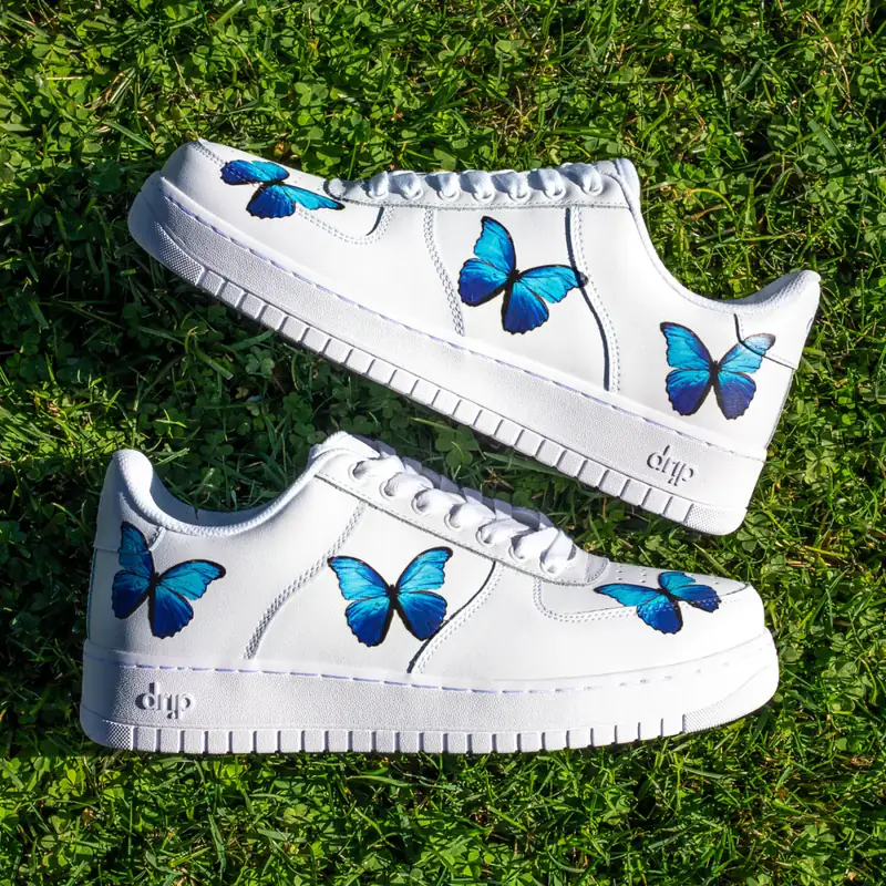 Product photograph of custom white sneakers with blue butterfly artwork, staged in bright grass