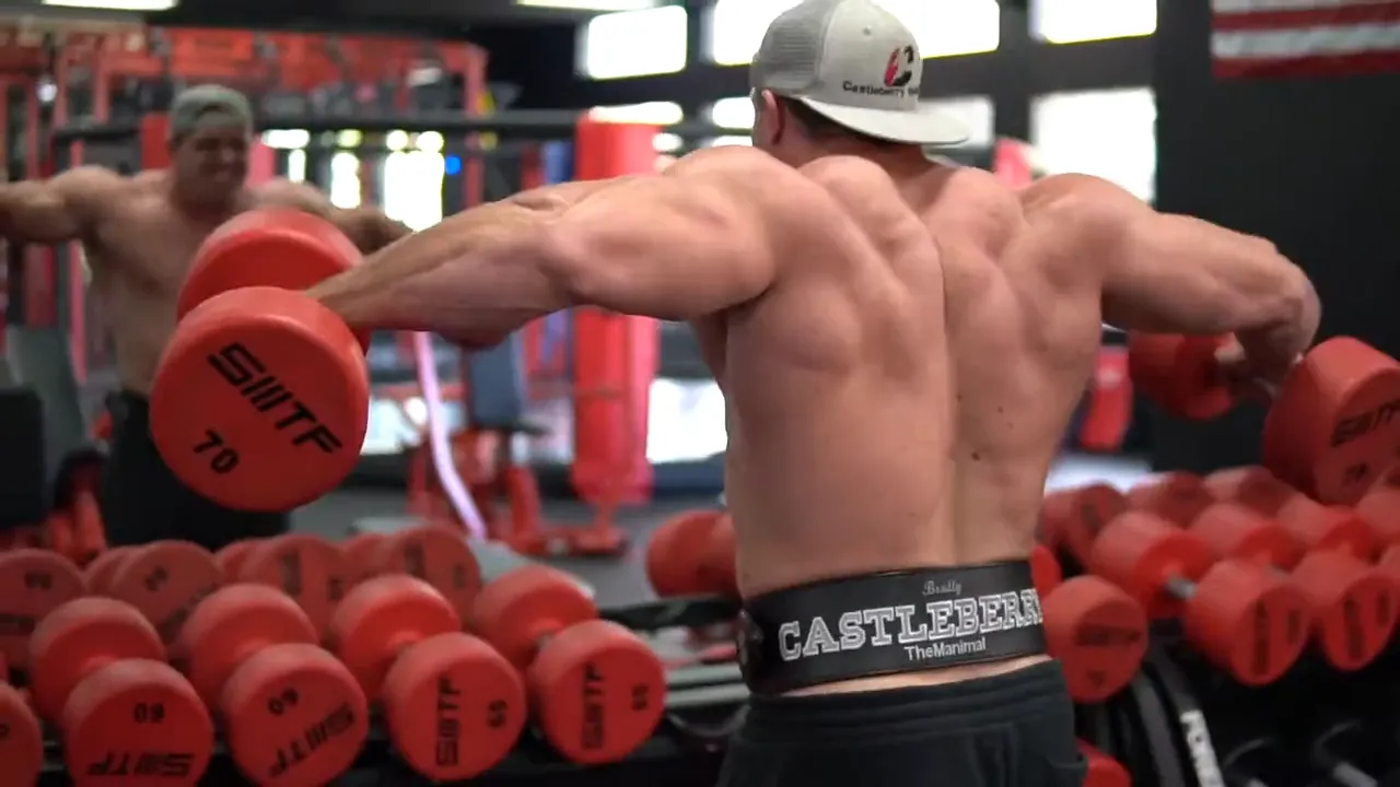 Lifter in a cap pressing a red dumbbell overhead beside a loaded gym rack
